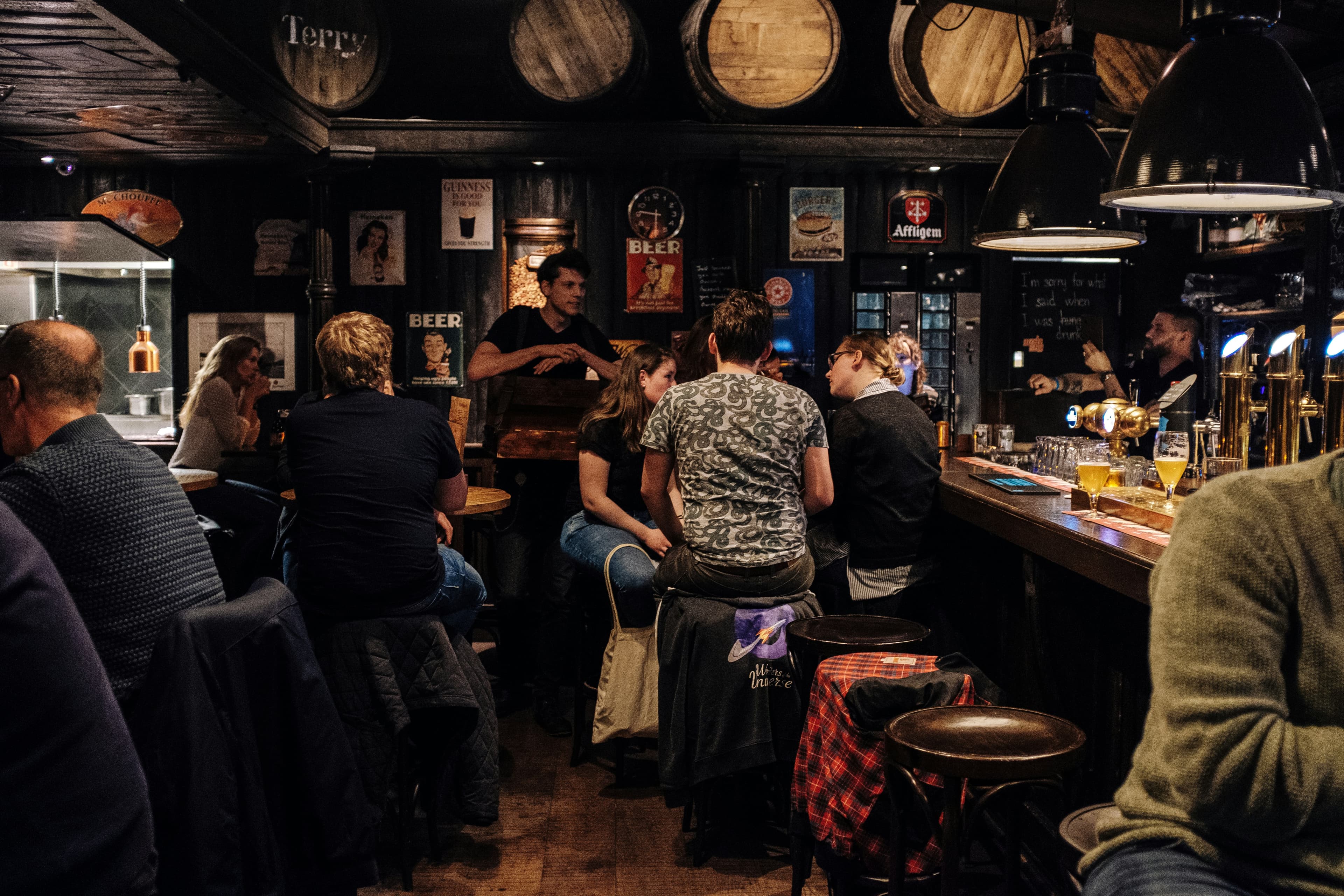 Busy wood-paneled pub with people chatting at the bar and tables beneath wooden barrels