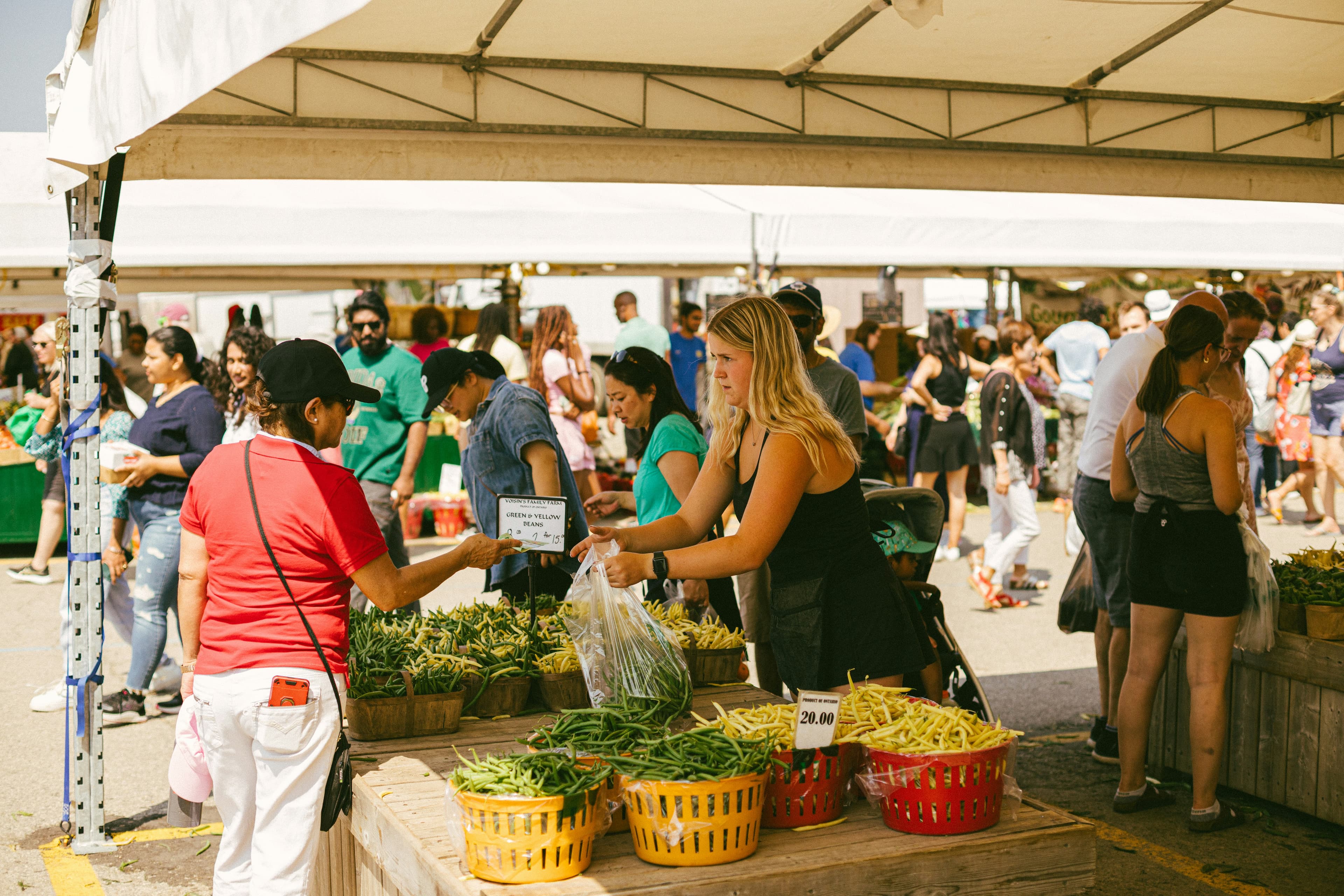 Shoppers browsing baskets of fresh produce under white tents at a busy outdoor market
